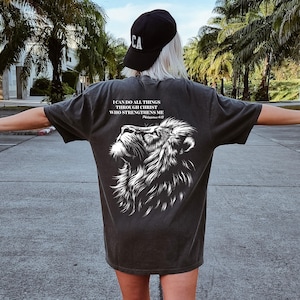 May include: Dark gray t-shirt featuring a detailed white lion graphic and the text "I CAN DO ALL THINGS THROUGH CHRIST WHO STRENGTHENS ME". The shirt is worn with a black baseball cap.