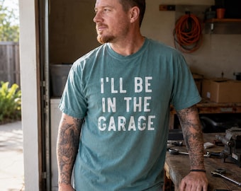 May include: A teal short-sleeved crew neck t-shirt featuring the white distressed text "I'LL BE IN THE GARAGE" printed across the chest. The shirt is made of a soft, textured fabric and has a standard fit with a ribbed collar and hemmed sleeves.