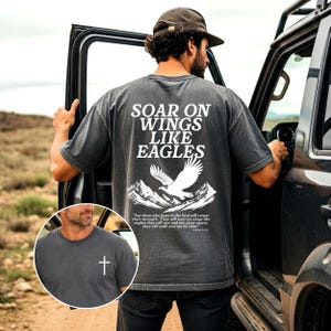 May include: Dark grey t-shirt with the text "SOAR ON WINGS LIKE EAGLES" and an eagle graphic. The shirt also features a small white cross on the chest. The back of the shirt has a mountain range and a bible verse.