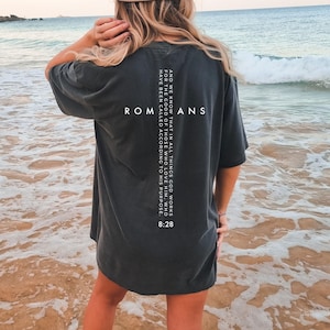May include: Dark gray oversized t-shirt with the text "ROMANS" and a Bible verse printed on the back. The verse reads, "AND WE KNOW THAT IN ALL THINGS GOD WORKS FOR THE GOOD OF THOSE WHO LOVE HIM, WHO HAVE BEEN CALLED ACCORDING TO HIS PURPOSE. 8:28."
