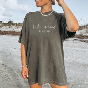 Peut inclure: Un t-shirt vert olive avec les mots "be transformed" et "Romans 12:2" imprimés en écriture blanche. Le t-shirt a une coupe décontractée et est porté sur une plage.