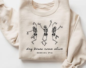 May include: A beige sweatshirt featuring a black graphic of three dancing skeletons. The text "dry bones come alive" and "EZEKIEL 37:5" are printed below the skeletons. The sweatshirt has a crew neck and long sleeves.