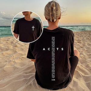 May include: Black t-shirt with a white cross on the front and the word "ACTS" on the back, along with a Bible verse. The shirt is worn on a beach with the ocean in the background.