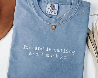 May include: A blue Comfort Colors t-shirt with the text "Iceland is calling and I must go." A gold necklace with a round pendant rests on the shirt. The shirt is folded and has a small tag at the neckline.