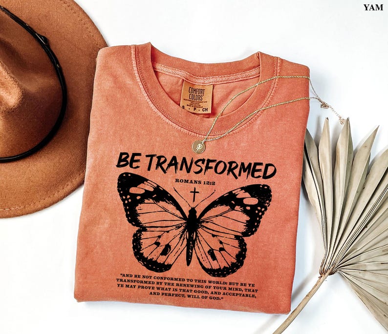 May include: A peach-colored t-shirt with a black butterfly graphic and the text "BE TRANSFORMED" with the Bible verse "Romans 12:2" and a quote from the Bible.