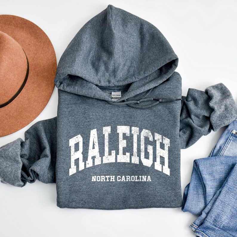 May include: A grey hoodie featuring the word "RALEIGH" in white, with "NORTH CAROLINA" underneath. The hoodie has a drawstring hood and long sleeves. A brown hat and blue jeans are next to the hoodie.