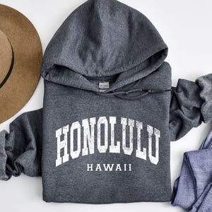 May include: A dark gray hooded sweatshirt with the word "HONOLULU" in white block letters arched across the chest, with "HAWAII" below. A brown hat and blue jeans are visible.