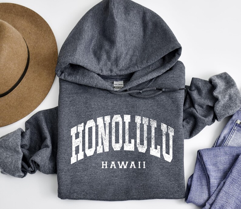 May include: A dark gray hooded sweatshirt with the word "HONOLULU" in white block letters arched across the chest, with "HAWAII" below. A brown hat and blue jeans are visible.