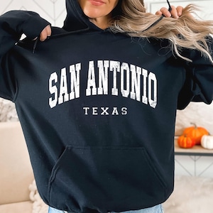 May include: Black hooded sweatshirt with the text "SAN ANTONIO TEXAS" in white, arched across the chest. The sweatshirt has a front pocket and a drawstring hood. The image is of a person wearing the sweatshirt.
