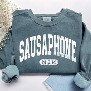 Peut inclure: Un sweat-shirt à col rond bleu ardoise avec le texte blanc vieilli SAUSAPHONE dans une police universitaire arquée au-dessus du mot MOM encadré par une bordure rectangulaire arrondie. Le vêtement porte une étiquette Comfort Colors au col.