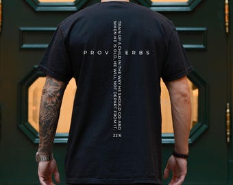 May include: A black short-sleeved t-shirt featuring white text on the back. The design includes the word PROVERBS horizontally and a vertical cross shape formed by the text: TRAIN UP A CHILD IN THE WAY HE SHOULD GO: AND WHEN HE IS OLD, HE WILL NOT DEPART FROM IT. 22:6.
