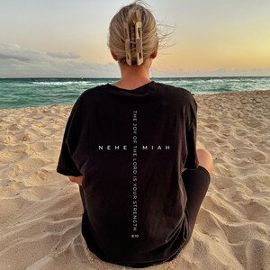 May include: A person wearing a black t-shirt with the text "THE JOY OF THE LORD IS YOUR STRENGTH" and "NEHEMIAH 8:10" printed in white. The person is sitting on a sandy beach, facing the ocean at sunset. A light-colored hair clip is visible.