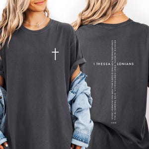 May include: Dark gray t-shirt with a white cross on the front. The back of the shirt features the text "1 Thessalonians" and a vertical list of inspirational text. The shirt has short sleeves and a relaxed fit.