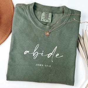May include: An olive green short-sleeve t-shirt with a crew neckline. The shirt features the white cursive text "abide" printed above the white text "JOHN 15:4". A gold-toned necklace with a small circular pendant rests on the fabric.