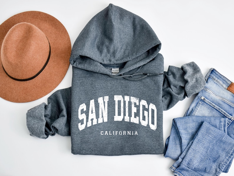 May include: A gray San Diego hoodie with white lettering and the word "California" underneath. A brown hat and blue jeans are next to the hoodie. The hoodie has a drawstring and a hood.