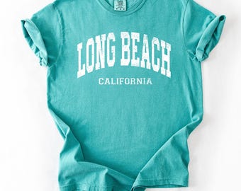 May include: A turquoise t-shirt with rolled-up sleeves. The shirt displays the text "LONG BEACH CALIFORNIA" in white, arched across the front. The t-shirt is made of a soft material, suitable for casual wear.