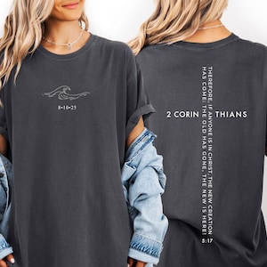 May include: Charcoal grey t-shirt with a minimalist wave design and the numbers 8-10-25 printed on the front. The back of the shirt features the text "2 Corinthians" and a vertical quote from the Bible, "Therefore, if anyone is in Christ, the new creation has come: The old has gone, the new is here! 5:17."