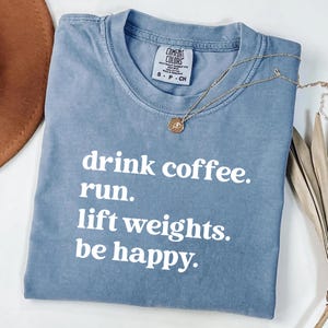 May include: A blue Comfort Colors t-shirt with the text "drink coffee. run. lift weights. be happy." in white. A gold necklace with a round pendant is draped across the shirt.