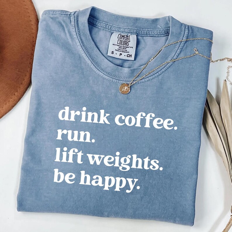 May include: A blue Comfort Colors t-shirt with the text "drink coffee. run. lift weights. be happy." in white. A gold necklace with a round pendant is draped across the shirt.