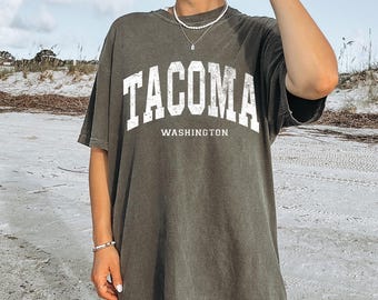 May include: Dark olive green t-shirt with the word "TACOMA" in large white block letters above "WASHINGTON". The shirt has a vintage, worn look and a relaxed fit.