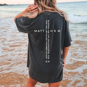 May include: Dark grey t-shirt with the text "MATTHEW" and a Bible verse printed vertically on the back. The verse reads "BLESSED ARE THE PEACEMAKERS, FOR THEY WILL BE CALLED CHILDREN OF GOD. 5:9". The shirt is worn on a beach.