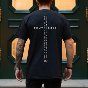 Peut inclure: T-shirt noir avec le mot "PROVERBES" et le texte "LES MÉCHANTS FUIENT QUOIQUE PERSONNE NE LES POURSUIVE, MAIS LES JUSTES SONT AUDACIEUX COMME UN LION. 28:1" imprimé en blanc. Le t-shirt est en matière douce et confortable.