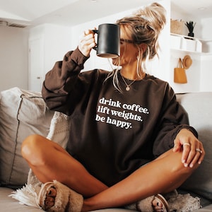 May include: A brown sweatshirt with the text "drink coffee. lift weights. be happy." in white. The person is holding a black mug. The sweatshirt is long-sleeved and has a crew neck.