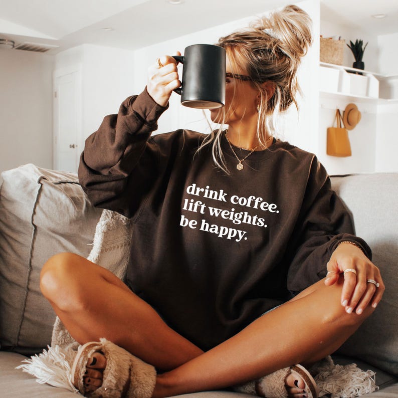 May include: A brown sweatshirt with the text "drink coffee. lift weights. be happy." in white. The person is holding a black mug. The sweatshirt is long-sleeved and has a crew neck.