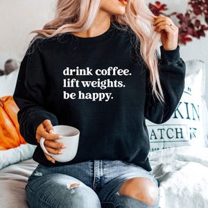 May include: Black sweatshirt with the text "drink coffee. lift weights. be happy." in white. The person is holding a white mug. The person is wearing blue jeans with a ripped knee.