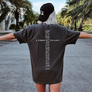 May include: A charcoal gray short-sleeve t-shirt featuring white text on the back. The text reads 2 CORINTHIANS 12:9 and includes a verse about grace and power arranged in a cross shape. A black baseball cap with the letters CA is also visible.