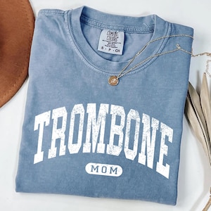 May include: A blue short-sleeved t-shirt with a distressed white graphic print that reads TROMBONE in an arched font and MOM inside a rounded rectangle below. The shirt features a Comfort Colors brand label at the neck and a size S-CH tag.
