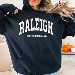 May include: Black hooded sweatshirt featuring the word "RALEIGH" in large, distressed white letters, and "NORTH CAROLINA" underneath. The sweatshirt has a front pocket and a drawstring hood. Pumpkins and a decorative item are also visible.