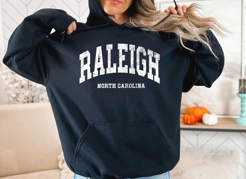 May include: Black hooded sweatshirt featuring the word "RALEIGH" in large, distressed white letters, and "NORTH CAROLINA" underneath. The sweatshirt has a front pocket and a drawstring hood. Pumpkins and a decorative item are also visible.