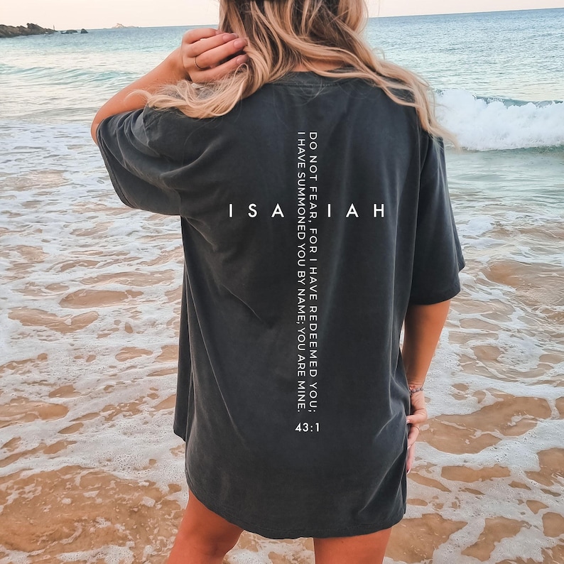 May include: Dark gray t-shirt with the word "ISAIAH" printed in white. The back of the shirt features the text "DO NOT FEAR, FOR I HAVE REDEEMED YOU; I HAVE SUMMONED YOU BY NAME; YOU ARE MINE. 43:1" in white. The shirt is worn on a beach.