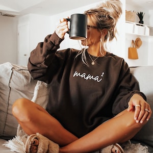 May include: A dark brown sweatshirt with the word "mama²" in white script. The person is holding a black mug. The sweatshirt is long-sleeved and appears to be made of a soft material. The person is sitting on a gray couch.