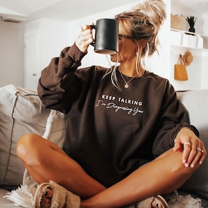 May include: A brown sweatshirt with the text "KEEP TALKING I'm Diagnosing You" in white script. The sweatshirt is worn with a black mug. The image is taken indoors.