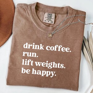 May include: A brown Comfort Colors t-shirt with the text "drink coffee. run. lift weights. be happy." A silver necklace with a small pendant is on the shirt.