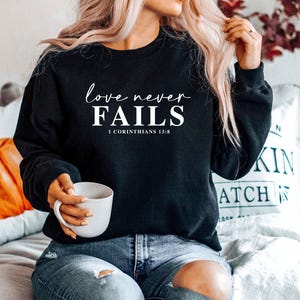 May include: Black sweatshirt with the text "love never FAILS" in a white script font, with "1 Corinthians 13:8" below. The sweatshirt is paired with blue jeans. A white mug is held in the hand.
