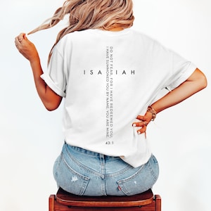 May include: White t-shirt with the word "Isaiah" and the text "Do not fear, for I have redeemed you; I have summoned you by name; you are mine. 43:1" printed in black. The shirt is worn by a person wearing blue jeans.