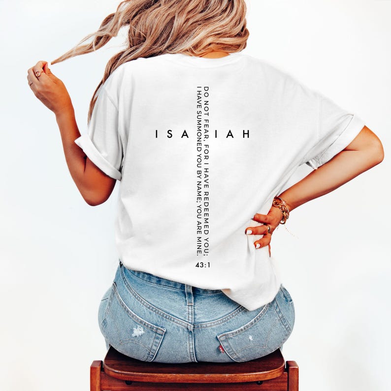 May include: White t-shirt with the word "Isaiah" and the text "Do not fear, for I have redeemed you; I have summoned you by name; you are mine. 43:1" printed in black. The shirt is worn by a person wearing blue jeans.