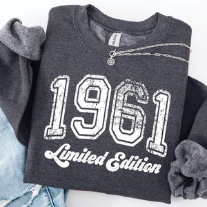 May include: A charcoal grey sweatshirt featuring the year "1961" in large, distressed white lettering, with "Limited Edition" below. A silver necklace with a sunburst pendant is draped across the garment. The sweatshirt is folded on a light surface.