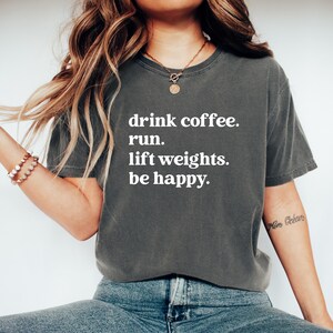 May include: Dark gray t-shirt with the text "drink coffee. run. lift weights. be happy." in white. The shirt is a relaxed fit and has short sleeves. The model is wearing blue jeans.