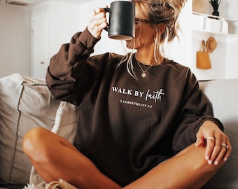 May include: A brown sweatshirt with the text "WALK BY faith" and "2 Corinthians 5:7" in white. The person is holding a black mug. The sweatshirt is long-sleeved and the person is wearing rings and a necklace.