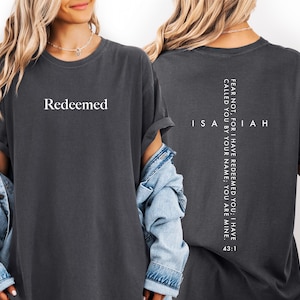 May include: Dark gray t-shirt with the word "Redeemed" in white on the front. The back of the shirt features a vertical cross design with the text "FEAR NOT, FOR I HAVE REDEEMED YOU; I HAVE CALLED YOU BY YOUR NAME; YOU ARE MINE. ISAIAH 43:1."