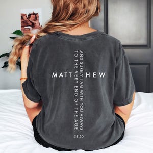 May include: Dark gray t-shirt with white text forming a cross shape. The word "MATTHEW" is centered horizontally, with the text "AND SURELY I AM WITH YOU ALWAYS, TO THE VERY END OF THE AGE. 28:20" vertically aligned.
