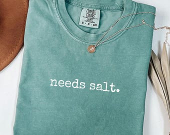 May include: A seafoam green Comfort Colors t-shirt with the phrase "needs salt." printed in white. A silver necklace with a small pendant rests on the shirt. The shirt is folded neatly.