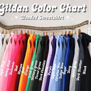 May include: A color chart for Gildan hooded sweatshirts, displaying a variety of colors including white, pink, red, orange, green, blue, gray, and black. The sweatshirts are hanging on a wooden rod, with the color names labeled below each garment.