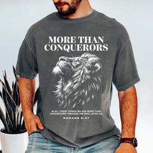 May include: Gray t-shirt with the text "MORE THAN CONQUERORS" above a detailed white lion illustration. Below the lion is the text "IN ALL THESE THINGS WE ARE MORE THAN CONQUERORS THROUGH HIM WHO LOVED US. ROMANS 8:37."