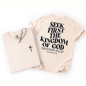 May include: Two cream-coloured t-shirts with black text and a silver chain necklace. One shirt has a black cross, the other has "SEEK FIRST THE KINGDOM OF GOD" printed on the back, with a Bible verse reference.