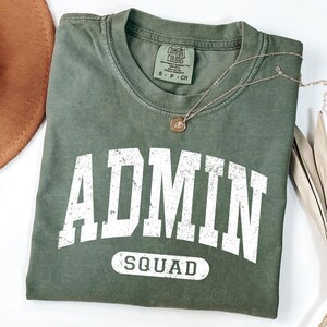 May include: Olive green t-shirt with the word "ADMIN" in large white letters and "SQUAD" underneath. A gold necklace with a small pendant rests on the shirt. The shirt is folded and has a tag that says "COMFORT COLORS".